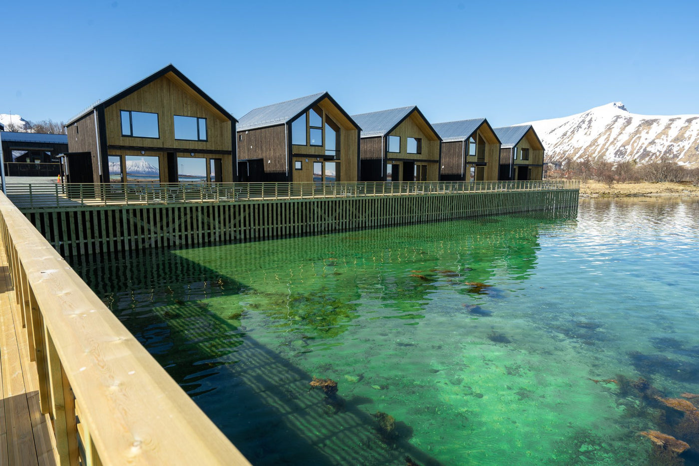 Trästugor pålar över turkost vatten, perfekt för sportfiskare. Träbrygga, snötäckta berg och blå himmel i bakgrunden.