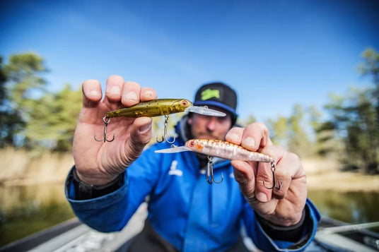 En sportfiskare i blå jacka visar två fiskedrag, ett grönt och ett brunt, mot en bakgrund av träd och blå himmel.