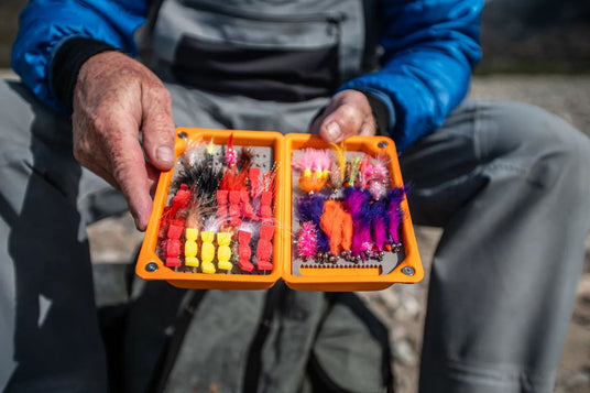 En person i friluftskläder håller en öppen orange ask med färgglada fiskeflugor och drag, redo för sportfiske eller fiskeresa.