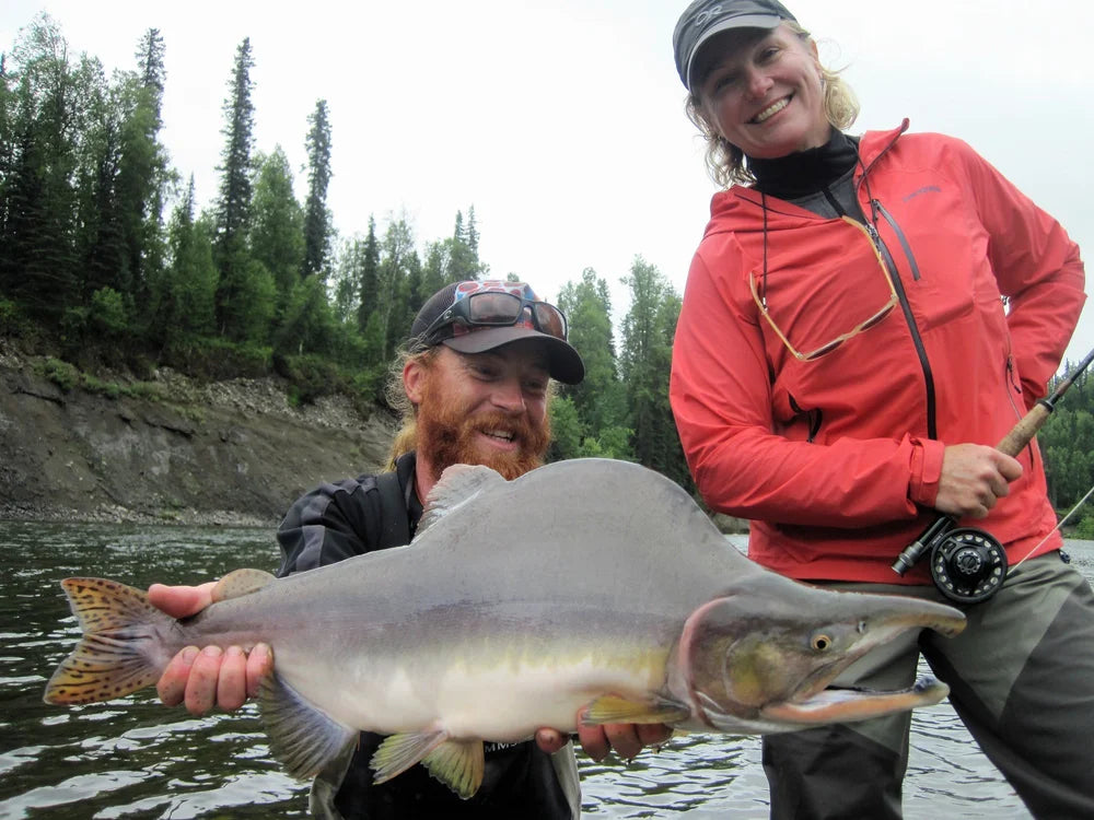 Leende sportfiskare visar stolt upp en stor lax fångad under en spännande fiskeresa vid en naturskön, trädklädd älv.