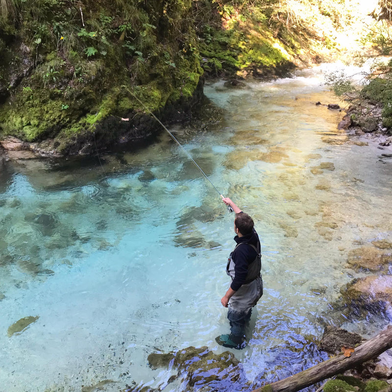 Sportfiskare i vadarbyxor flugfiskar i klar grund älv bland mossiga stenar och grönska, perfekt för fiskeresa.