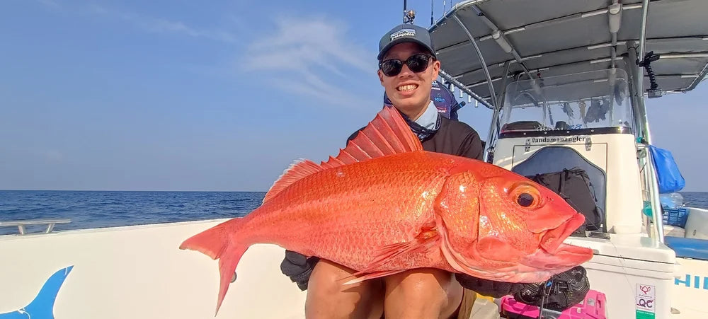 En person med solglasögon och keps ler på en båt, håller en stor röd fisk under en fiskeresa med hav och blå himmel i bakgrunden.