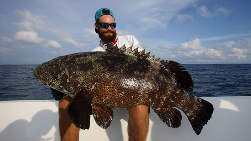 En man med solglasögon och bakåtvänd keps håller stolt en stor, prickig fisk på båt efter spännande fiskeresa till havs.