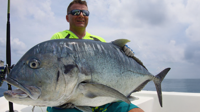 Leende man med solglasögon och handskar håller en stor silverfisk på båt under fiskeresa, hav och molnig himmel i bakgrunden.