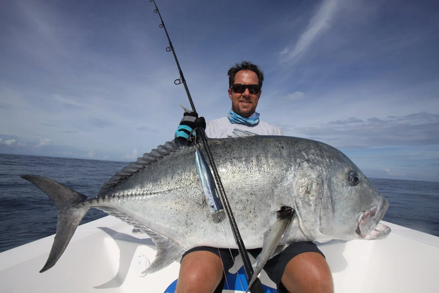 En sportfiskare med solglasögon och handskar sitter i en båt, håller ett stort silvrigt fiskefångst och spö mot havet och molnig himmel.