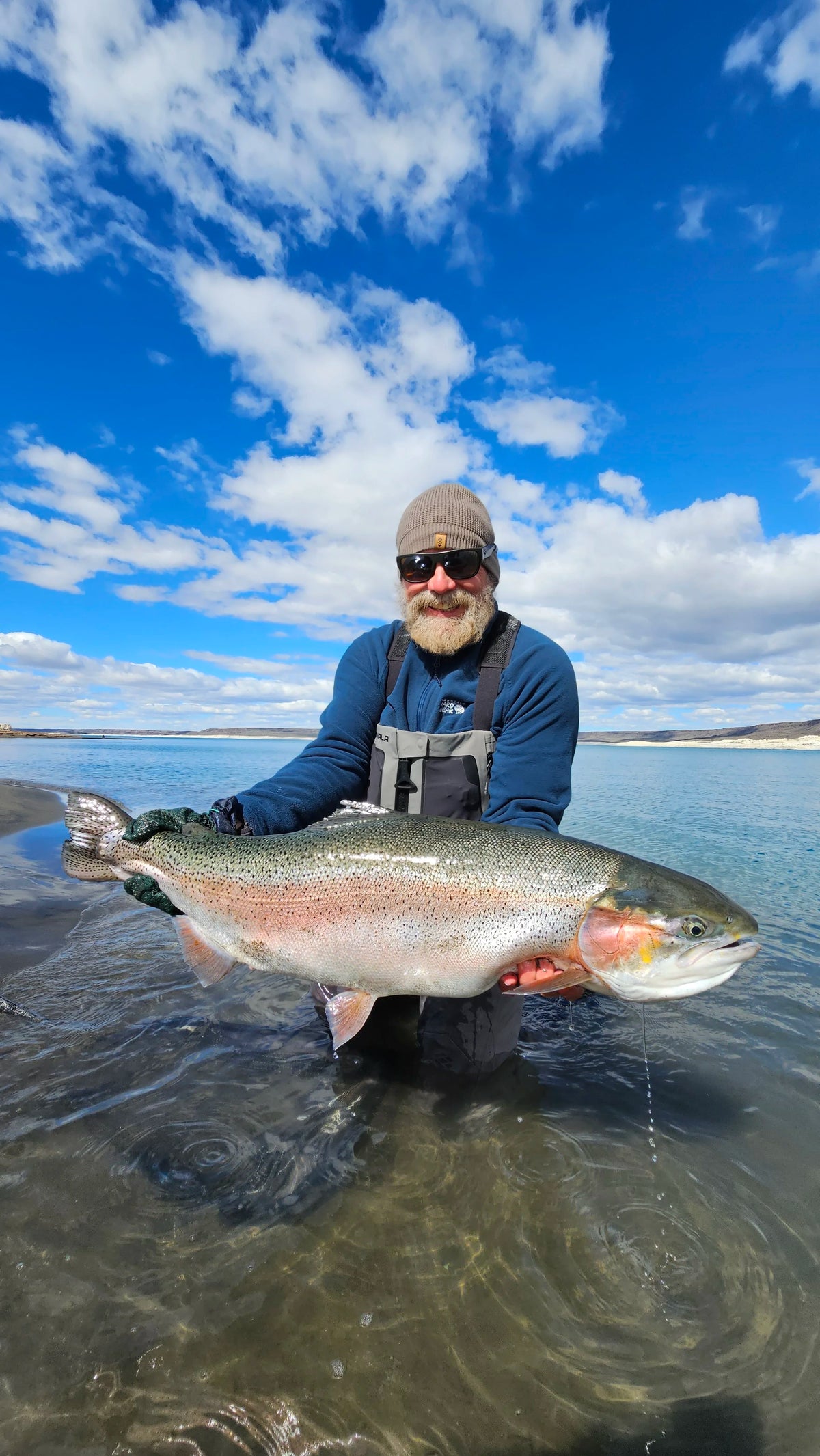 Skäggig man med hatt och solglasögon står i grunt vatten, ler och håller upp en stor färgglad fisk under en fiskeresa.