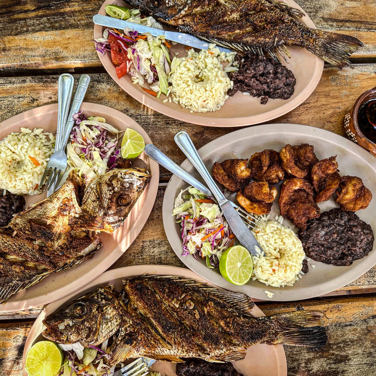Tre tallrikar med grillad hel fisk, ris, svarta bönor, sallad och lime samt en tallrik friterad fisk på rustikt träbord.