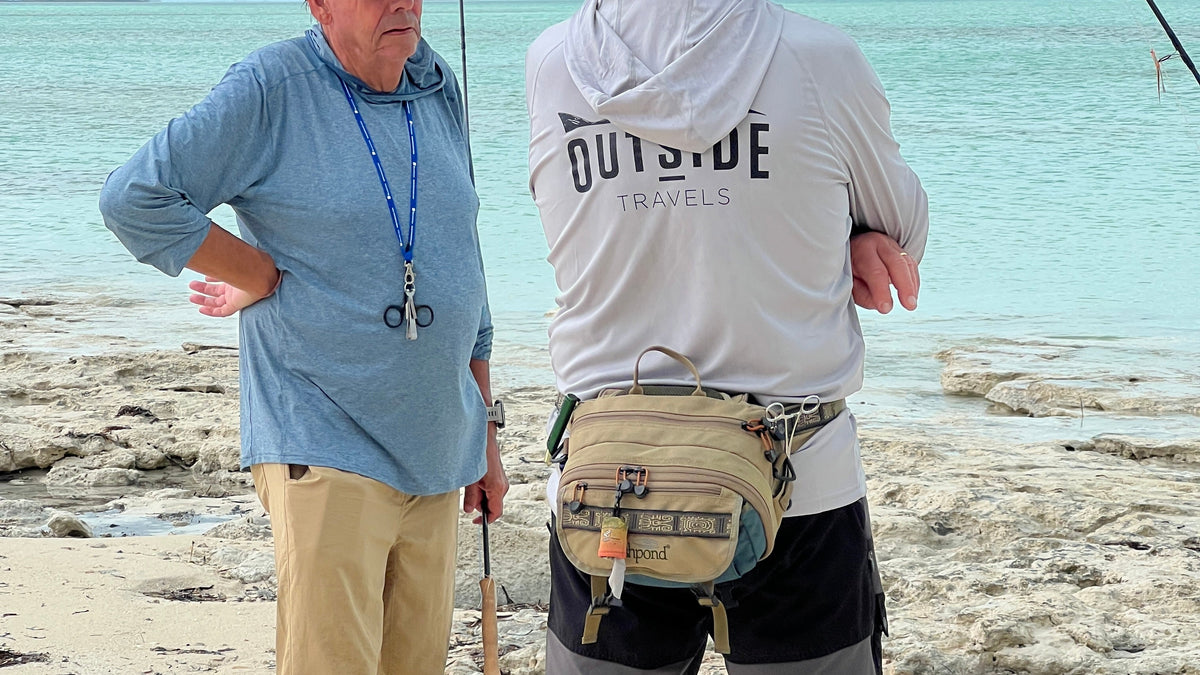 Två män på en klippig strand, fiskar tillsammans. En har blå skjorta, den andra OUTSIDE TRAVELS-hoodie och fiskeutrustning.