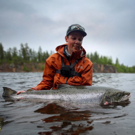 Sportfiskare med stor fångst i orange jacka vid skogskantat vatten, Fiskeresa Camp Onka Lappland.