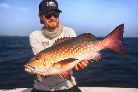 Sportfiskare med solglasögon, keps och buff visar stolt upp stor rödaktig fisk på båt under fiskeresa, hav och himmel i bakgrunden.