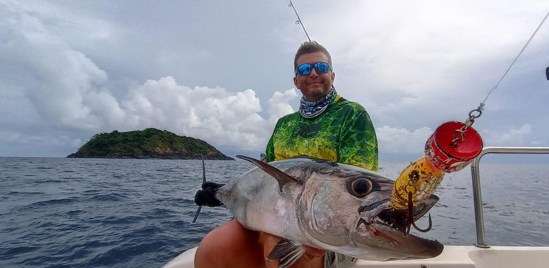 Sportfiskare med solglasögon och grön skjorta håller en stor fisk på båt, hav och grönskande ö i bakgrunden under molnig himmel.