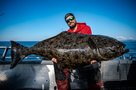 Man i röd hoodie och solglasögon håller upp en stor prickig fisk på båt under fiskeresa, blå himmel och hav syns bakom.
