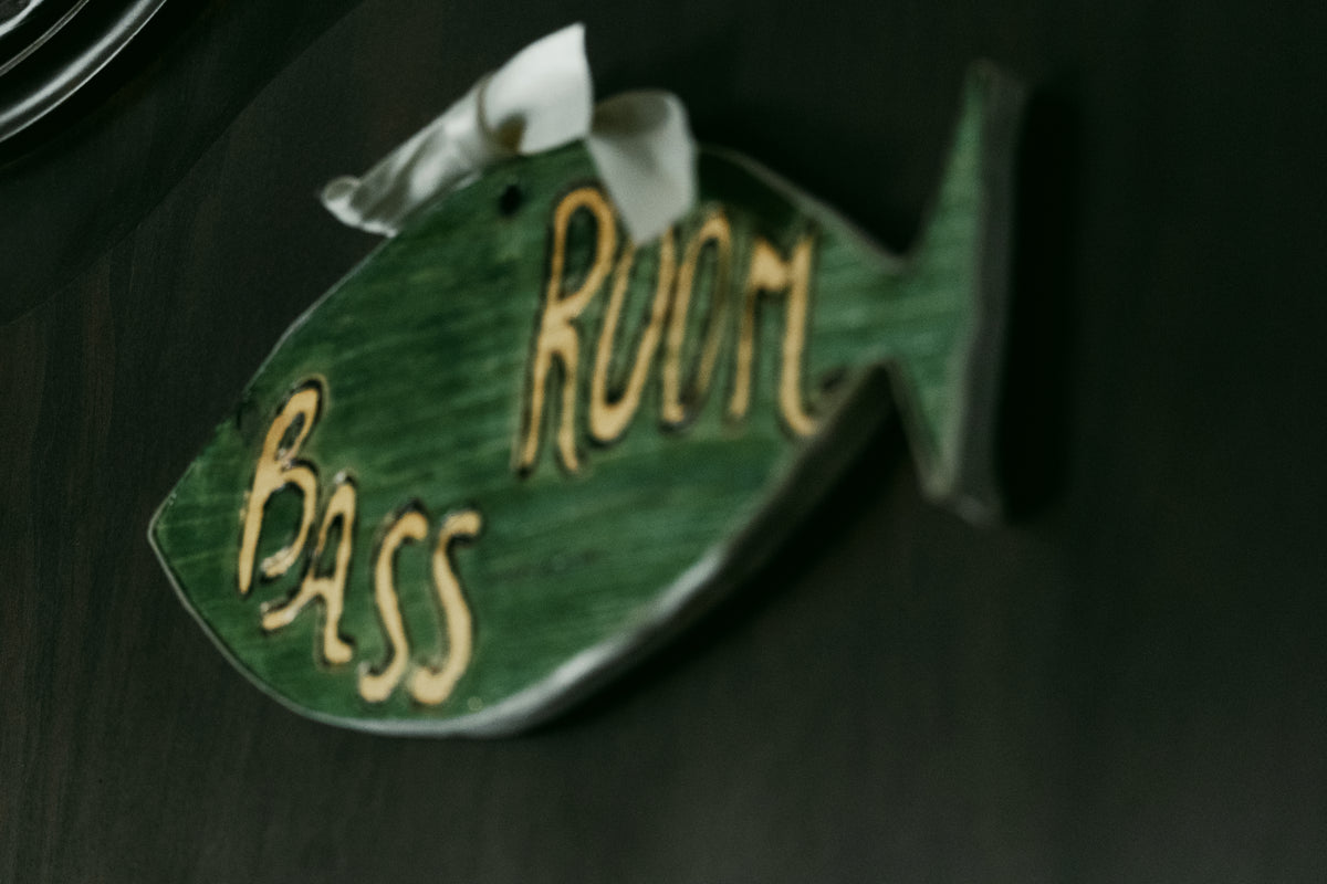 En grön fisformad skylt med texten Bass Room hänger på mörk vägg, dekorerad med vitt band - idealisk för sportfiskare.