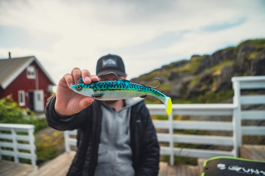 Sportfiskare i svart jacka och keps visar upp blå-gult fiskedrag. Röd stuga och klippor syns i bakgrunden.