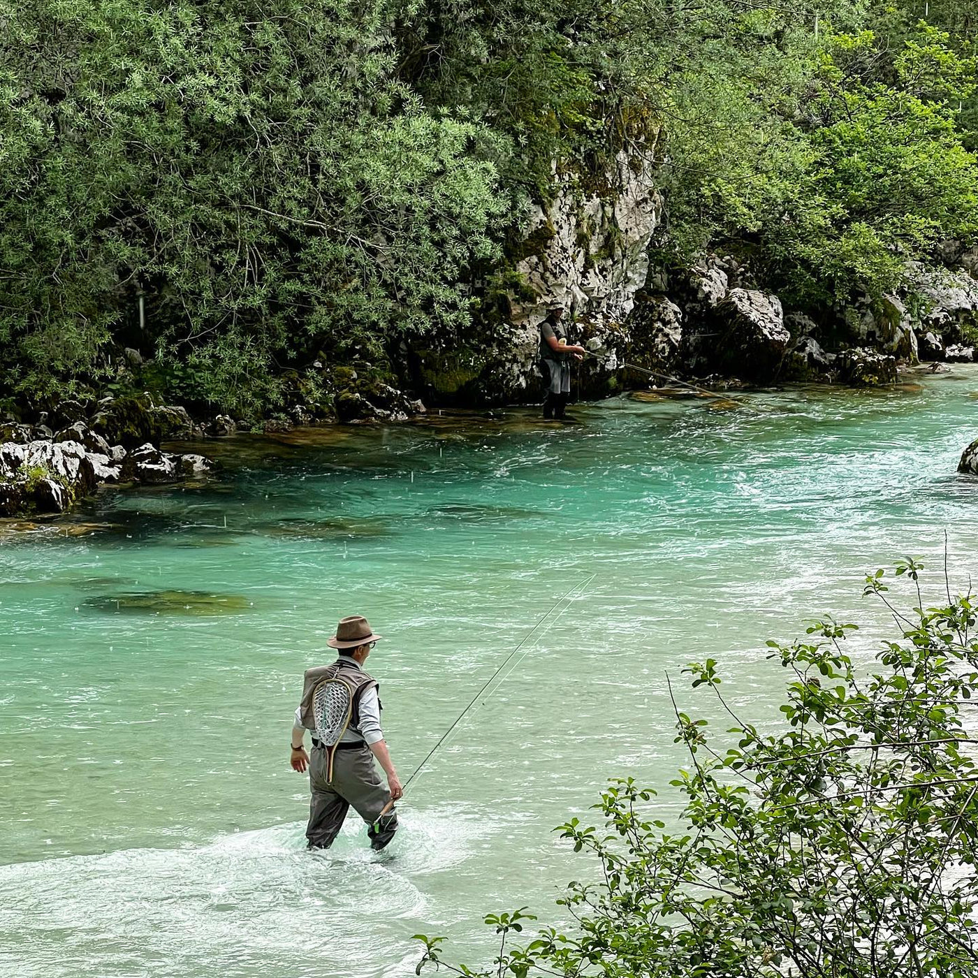 Två sportfiskare flugfiskar i klar, turkos älv omgivna av grönska och klippor under en minnesvärd fiskeresa.