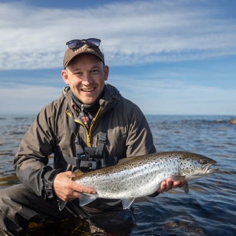 Leende sportfiskare i fiskeutrustning knäböjer i vattnet och visar stolt upp en stor prickig fisk på fiskeresa Gotland, Sverige.