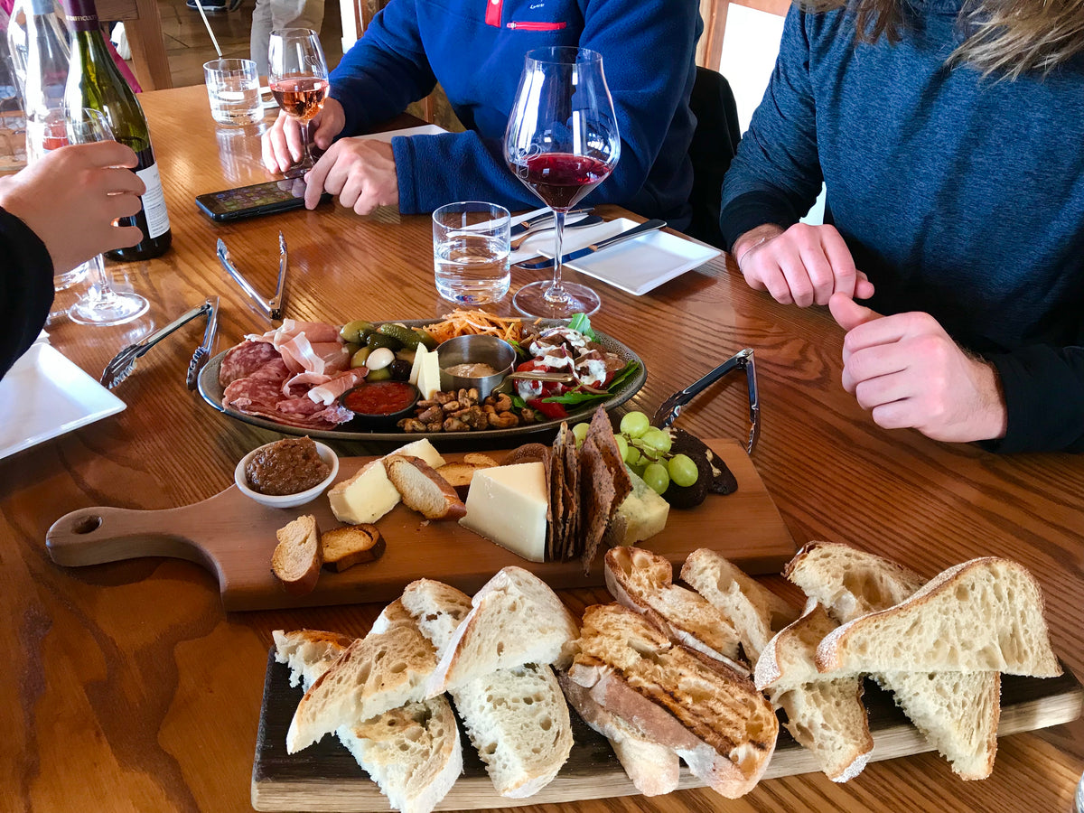 Träbord dukat med charkbricka, bröd och vin där tre sportfiskare från fiskeresa samlas; en häller upp vin, en använder mobil.