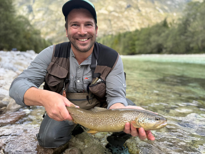 En leende sportfiskare i fiskeutrustning knäböjer vid en klar älv och håller upp en stor fisk, omgiven av fjäll och skog.