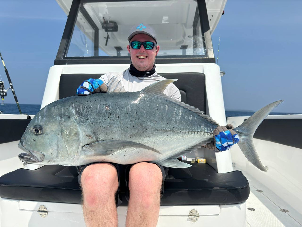 En leende sportfiskare med solglasögon, kepsar och handskar håller en stor fisk på båt under fiskeresa. Spön syns i bakgrunden.