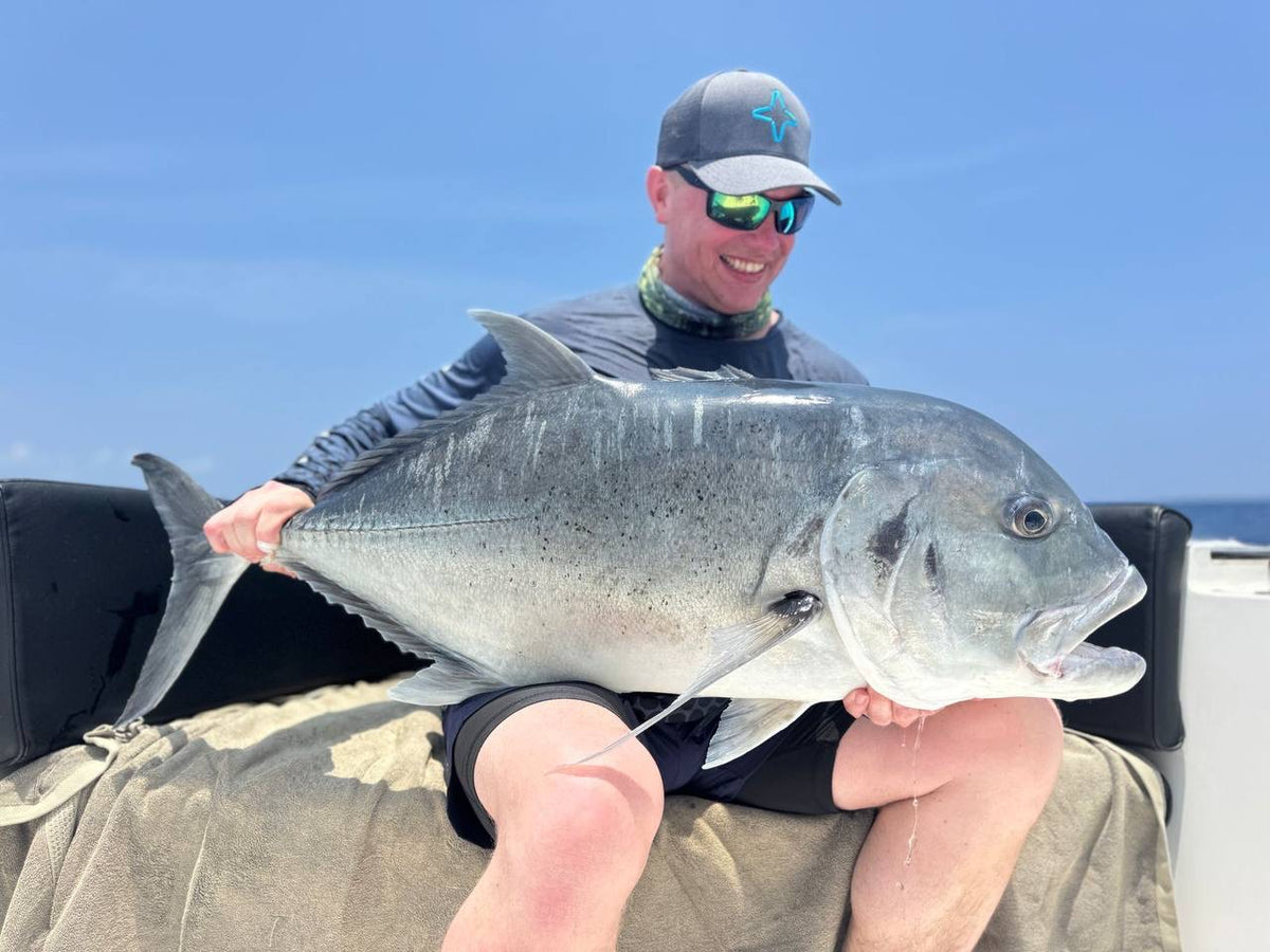 Sportfiskare med solglasögon, keps och buff sitter i båt, håller en stor silverfisk och ler mot havet under blå himmel.