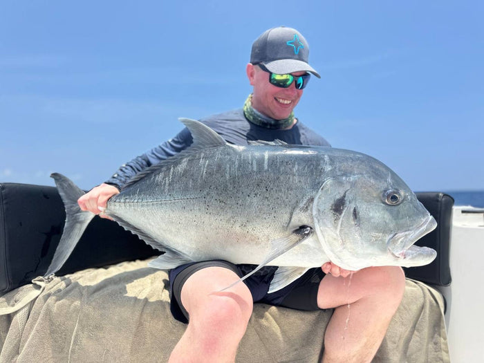 Sportfiskare med solglasögon, keps och buff sitter i båt, håller en stor silverfisk och ler mot havet under blå himmel.