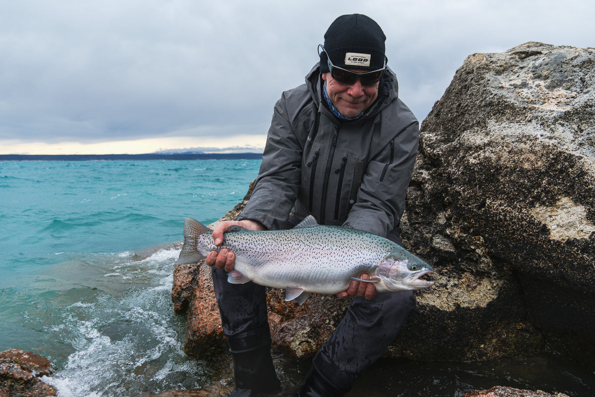 En sportfiskare i friluftskläder och solglasögonknäböjer vid en turkos sjö, håller stolt upp en stor fisk mot vågorna.