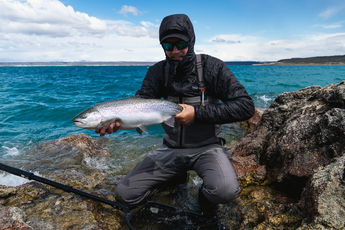 Sportfiskare i vattentäta kläder på stenig strand visar stolt upp stor fisk vid blått vatten under molnig himmel.