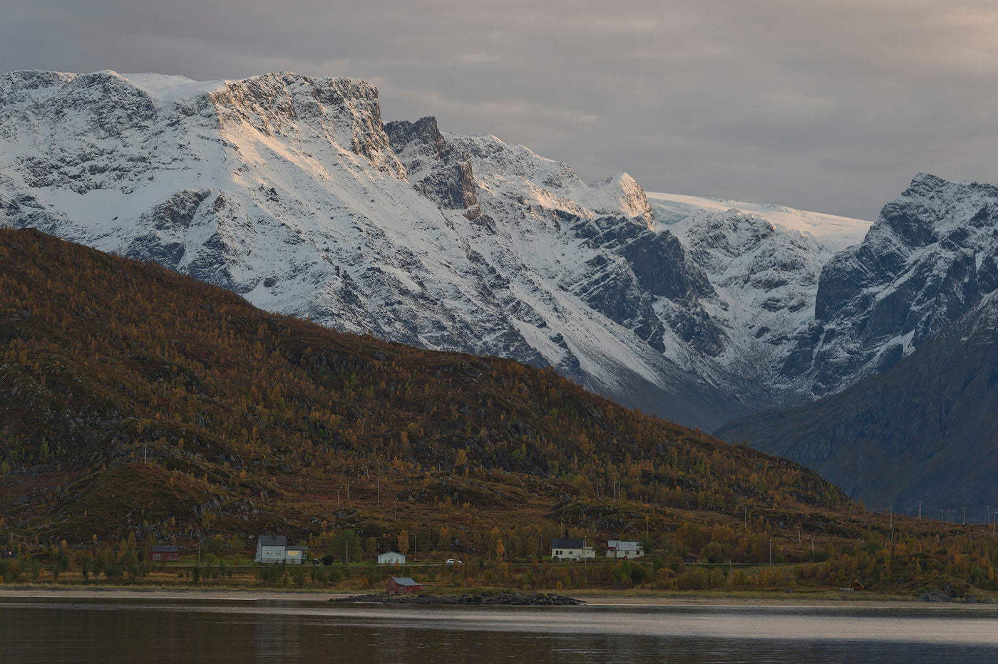 Snötäckta berg reser sig bakom skogsklädda kullar och hus. En sportfiskare njuter av fiske i lugnt vatten med vacker utsikt.