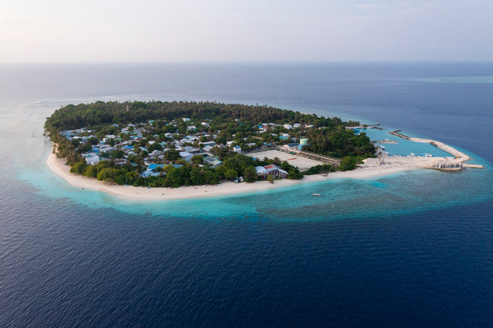 Flygbild av grönskande ö med byggnader och sandstränder, omgiven av turkost hav och korallrev - perfekt för sportfiske och fiskeresa.
