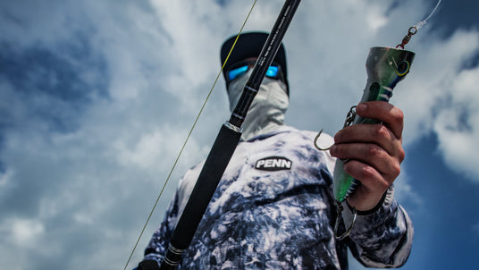 Sportfiskare med solglasögon, ansiktsmask och PENN-tröja håller fiskespö och stor wobbler under molnig himmel inför fiskeresan.