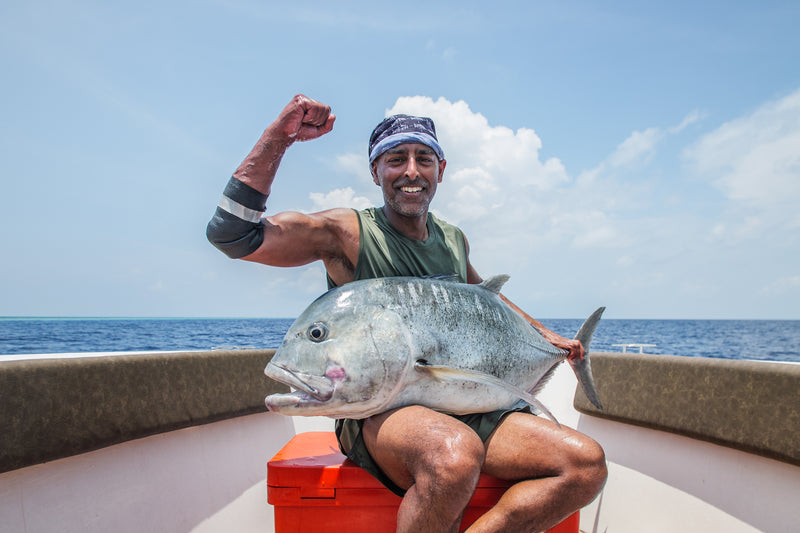 En leende sportfiskare på båt håller stor fisk, flexar armen, hav och blå himmel i bakgrunden-en idealisk fiskeresa.