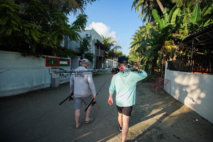 Två sportfiskare i fiskeutrustning går längs en sandstig med spön, omgivna av tropiska växter och vita hus under blå himmel.