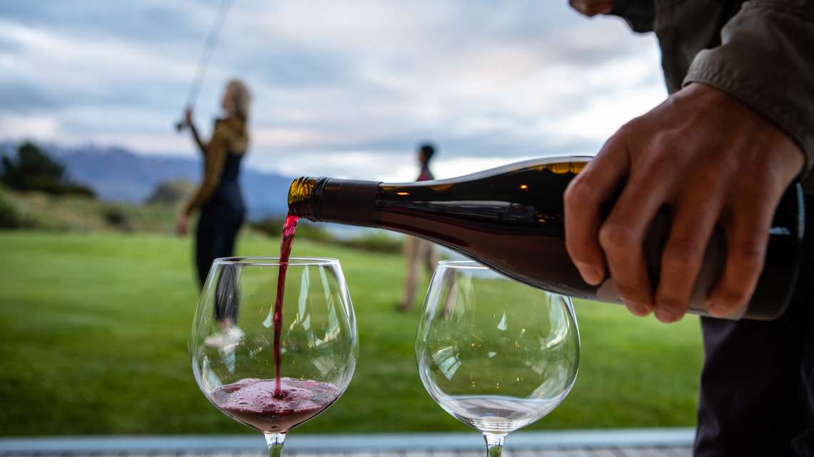 Närbild av hand som häller rött vin i glas utomhus, med två suddiga personer i bakgrunden efter en fiskeresa.