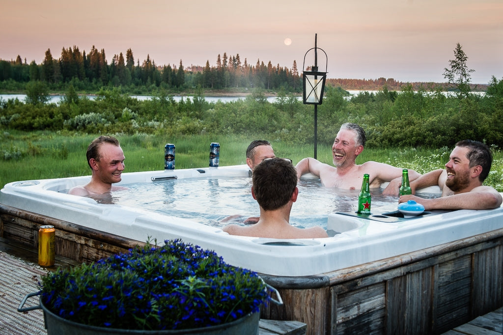 Fem män njuter i utomhusbubbelpool med öl vid sjön efter lyckad fiskeresa, omgivna av grönska och kvällssol.