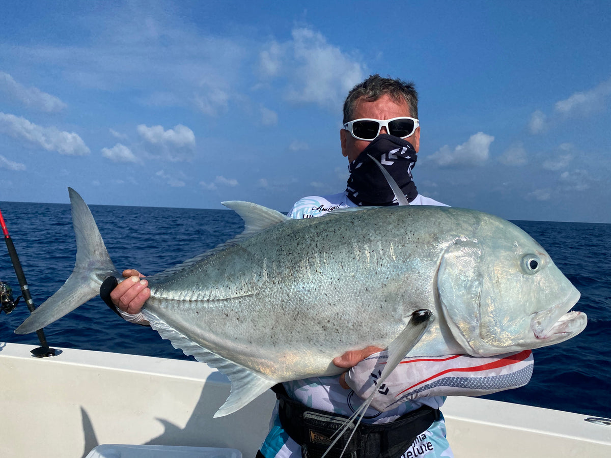 En man med solglasögon och ansiktsmask håller en stor silverfisk på en båt under blå himmel-drömresa för sportfiskare.