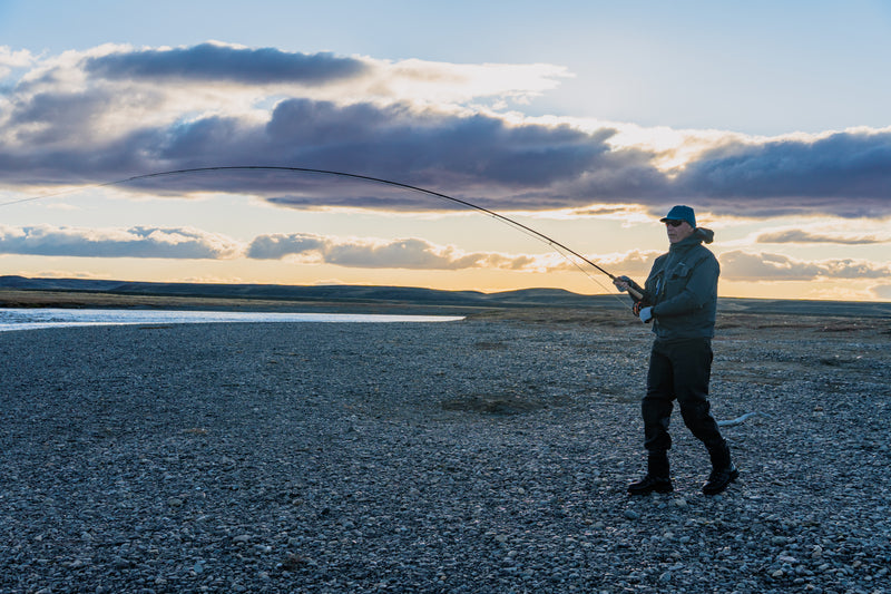 Sportfiskare i fiskeutrustning och solglasögon flugfiskar videnig kust i solnedgång-idealisk plats för fiskeresa.