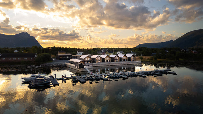 En rad trästugor vid vattnet, fiskebåtar i lugn marina, omgiven av berg och skog, speglar solnedgångens färger.