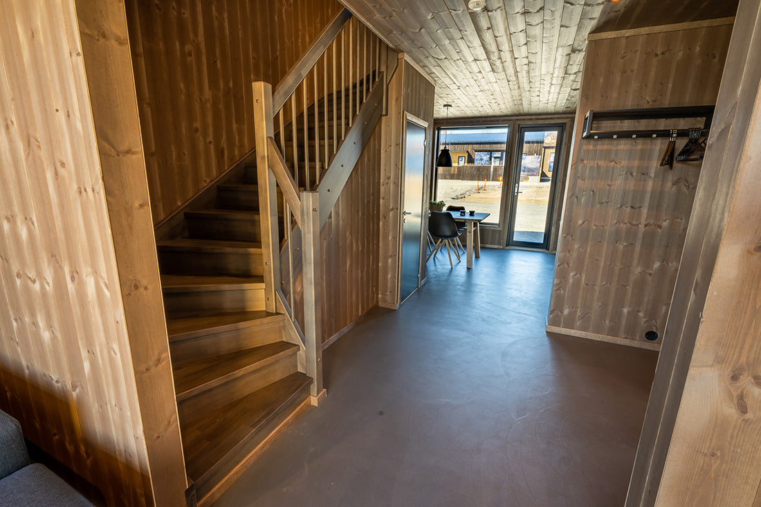 Interiör av timmerstuga med trappa till vänster, matbord för sportfiskare vid stora fönster och naturligt ljusflöde.