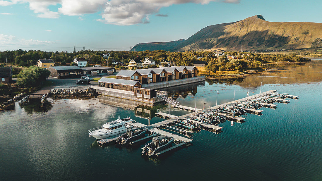 Flygvy över småbåtshamn där sportfiskare gör sig redo vid träbyggnader. Vatten, träd, hus och berg i bakgrunden.