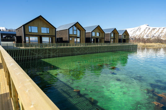 Trästugor pålar över turkost vatten, perfekt för sportfiskare. Träbrygga, snötäckta berg och blå himmel i bakgrunden.
