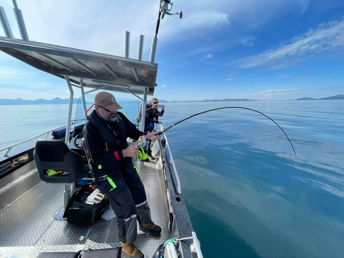 Två sportfiskare fiskar från båt på lugnt blått vatten. En person drillar fiskespö. Fjäll syns i bakgrunden.