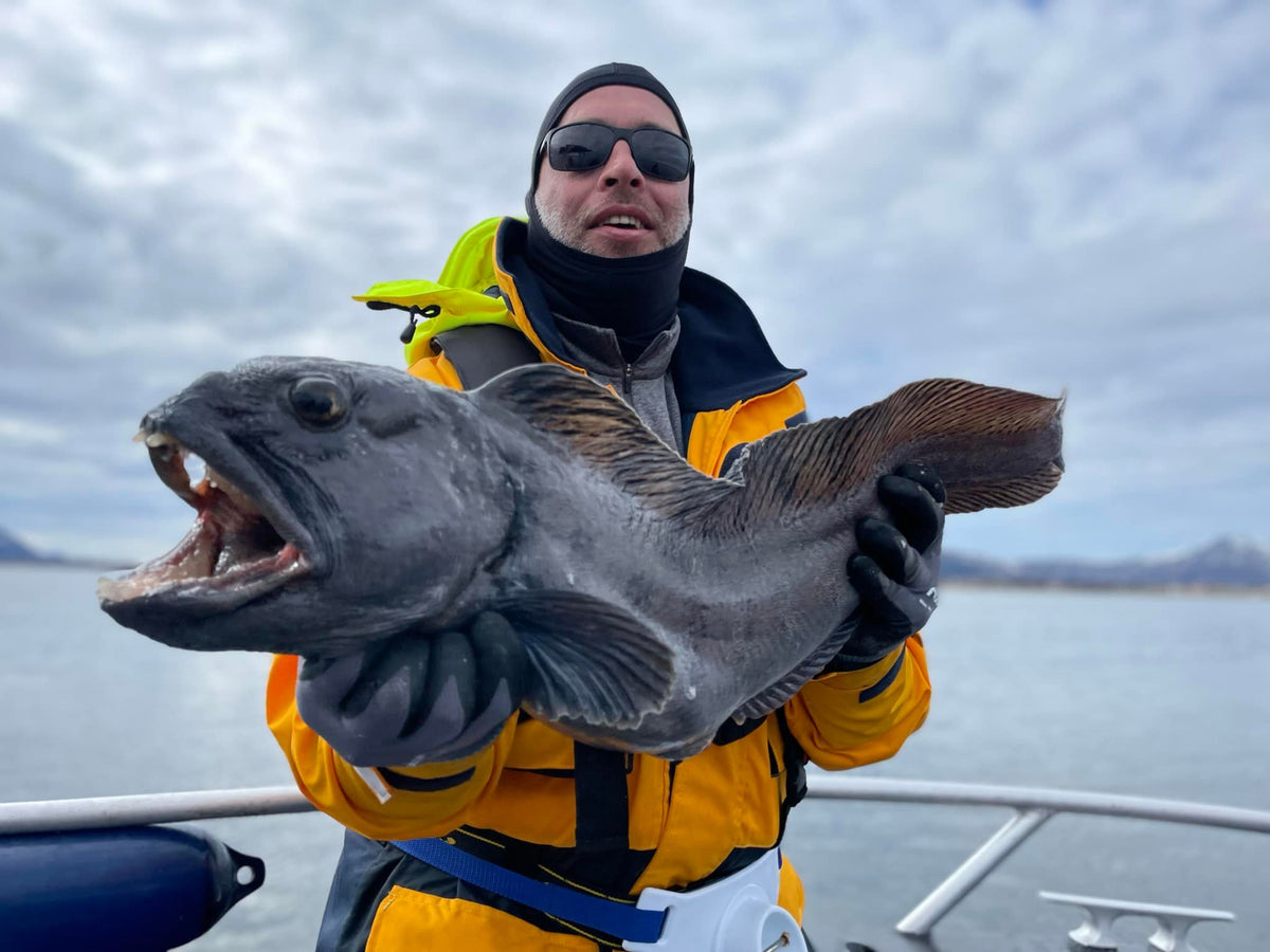 En sportfiskare i gul jacka och solglasögon visar upp en stor fisk ombord på en båt under fiskeresa, vatten och berg bakom.