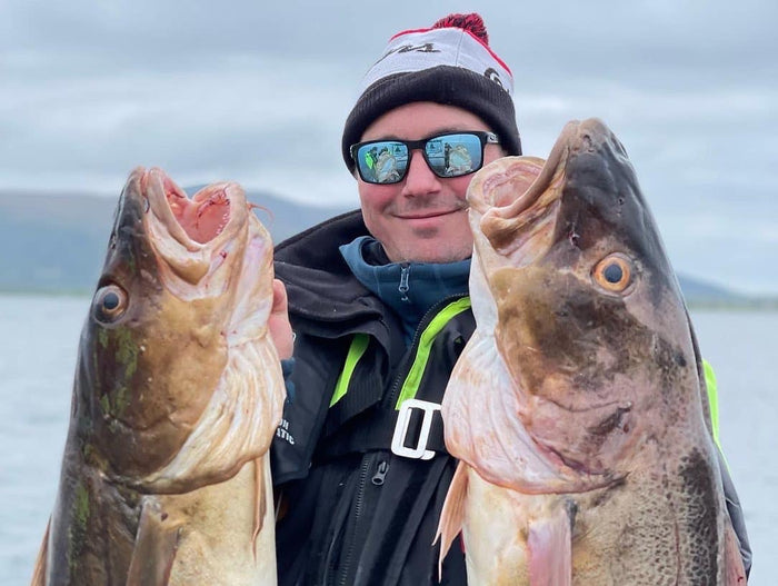 En leende sportfiskare med solglasögon, vintermössa och jacka håller stolt upp två stora fiskar vid vattnet under molnig himmel.