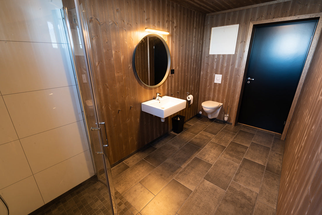 Modernt badrum med träpanel, rund spegel, vitt handfat, svart dörr, vägghängd toalett och dusch-idealisk efter fiskeresa.