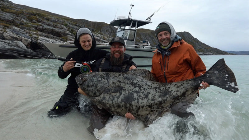 Tre glada sportfiskare i friluftskläder knäböjer i vattnet med en stor fisk. Båt och klippor syns under mulen himmel.