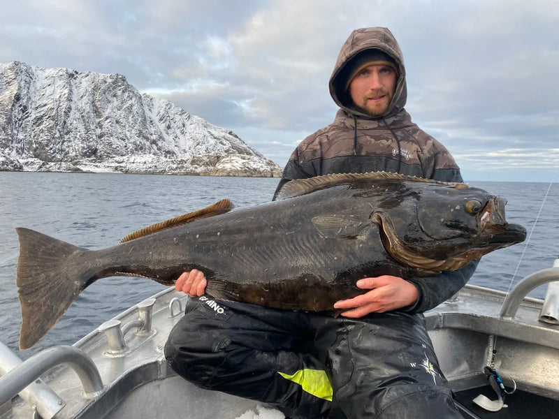 Sportfiskare i huva sitter på båt med stor mörk fisk, snötäckta fjäll och vatten under molnigt himmel, perfekt fiskeupplevelse.
