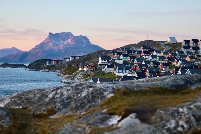 Färgglada hus vid stenig kust och lugnt vatten där sportfiskare samlas. Bergstopp i bakgrunden under mjuk pastellhimmel.