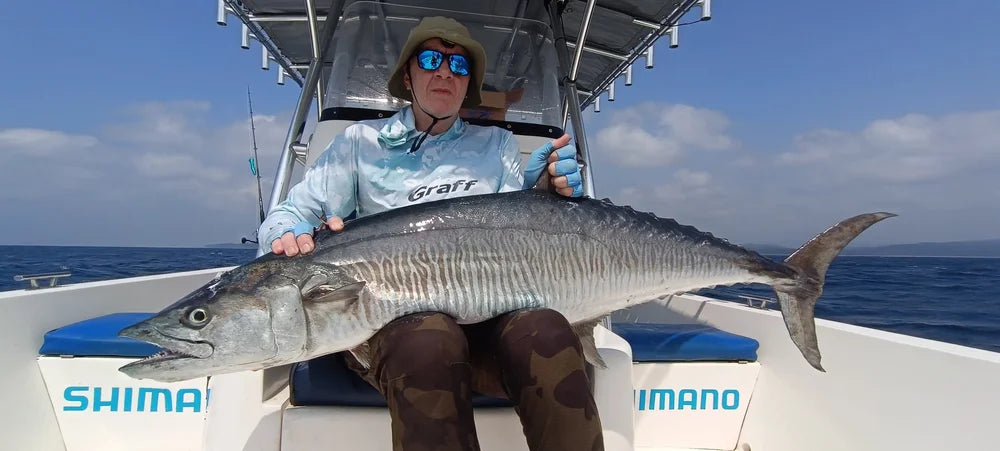 Sportfiskare med solglasögon och hatt håller en stor kungsmakrill på båt, hav och blå himmel i bakgrunden. Shimano-säten.