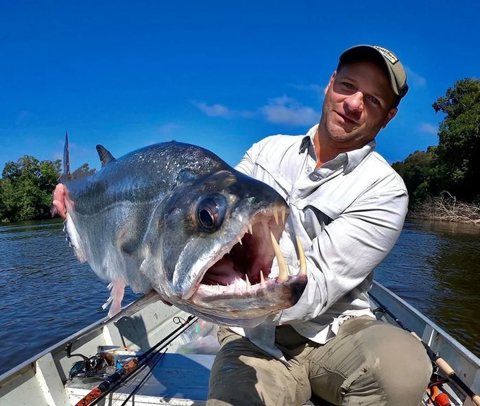 En sportfiskare i båt visar stolt en stor rovfisk under fiskeresan, med flod, grönska och blå himmel i bakgrunden.