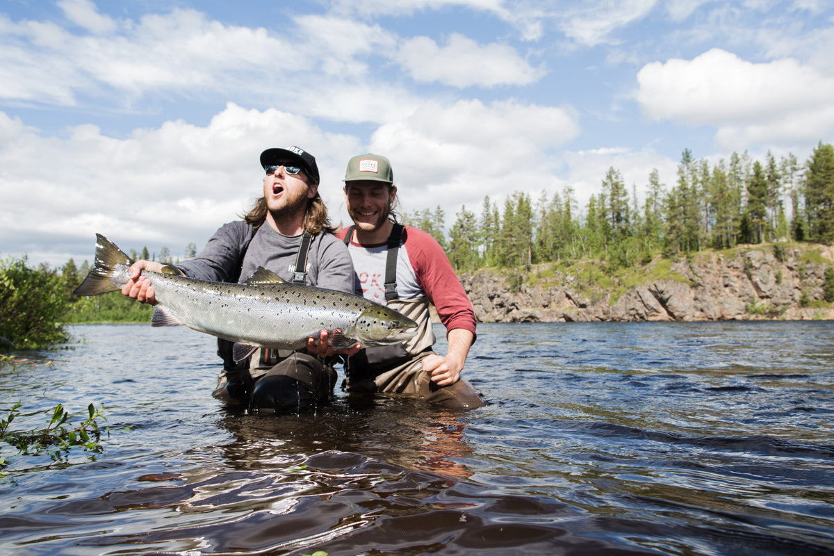 Två sportfiskare i vadare står i en älv, håller upp en stor fisk och firar en lyckad fiskeresa i naturskön omgivning.